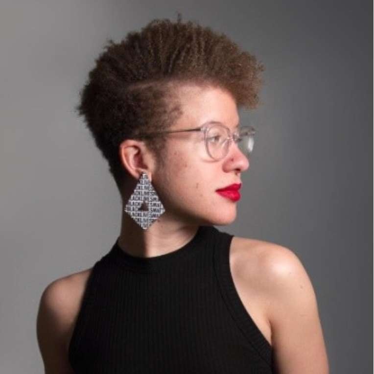 image of a lightskin femme with a short brown parted afro. They are wearing red lipstick, clear glasses, a black tank top, and a chandelier earring. They are looking over their left shoulder.