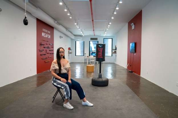 Session artist Caroline Garcia sitting in the gallery space. There is a punching bag, a wall of 3d printed weaponry, and monitors showing fight choreography. Image credit: Manuel Molina, courtesy of Recess