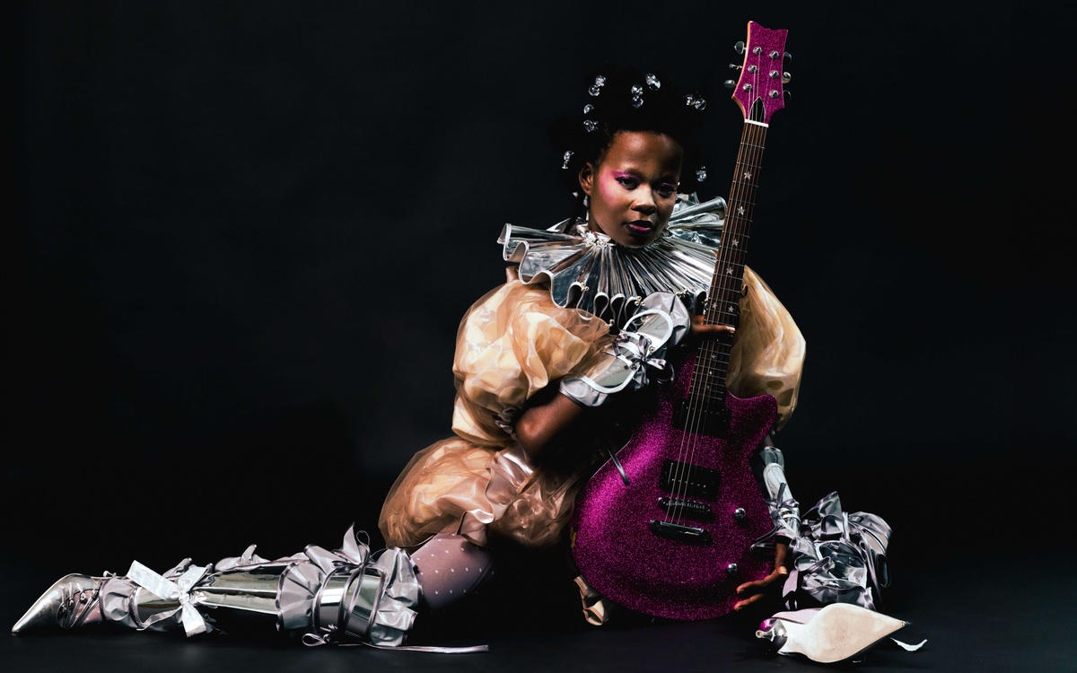 seated black femme in clown outfit straddling a purple guitar