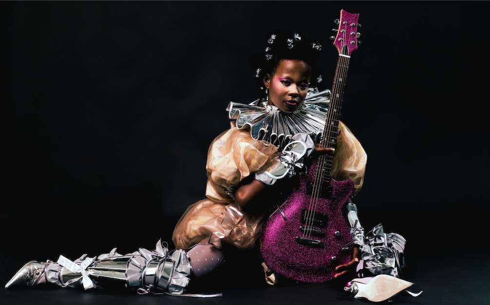 seated black femme in clown outfit straddling a purple guitar