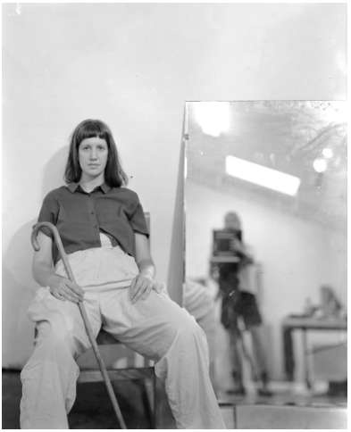 a black and white photo, by francie seidl chodosh, of anna roberts-gevalt. Anna, a white person with light hair with bangs and shoulder length hair, sits leaning against the wall. She stares directly at the camera, in a button up shirt, holding a cane in their hand. In the mirror, the photographer is partially glimpsed, blurry, behind a large camera.